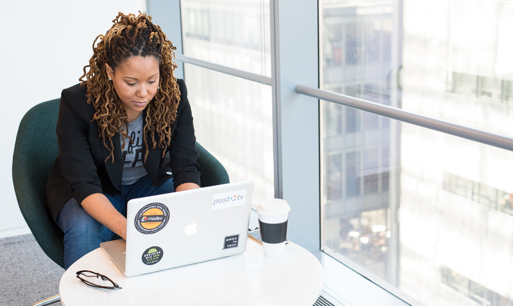 Photo of a brown skin person with long dreadlocks sitting beside a large window, typing on a laptop.